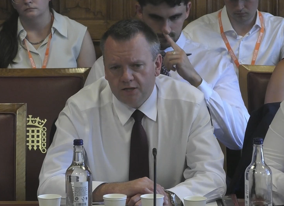 Minister Nick Thomas-Symonds in a white shirt and tie sitting at a table in front of the  Committee with a microphone and a bottle of water