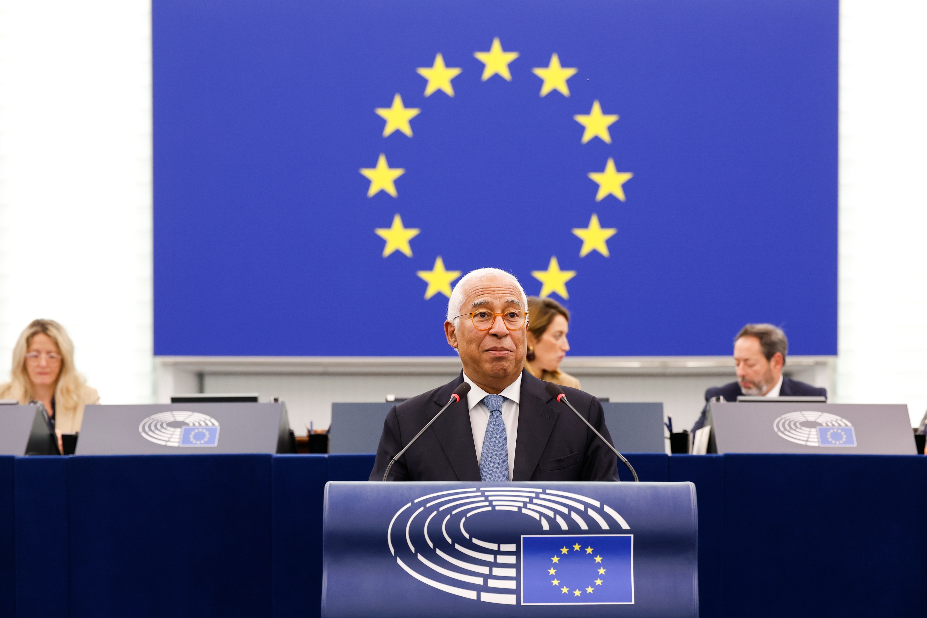 image shows the President of the European Council is António Costa addressing MEPs in the European Parliament. He is standing at a podium with the European Parliament logo, there is a blue banner behind him with gold stars in a circle.