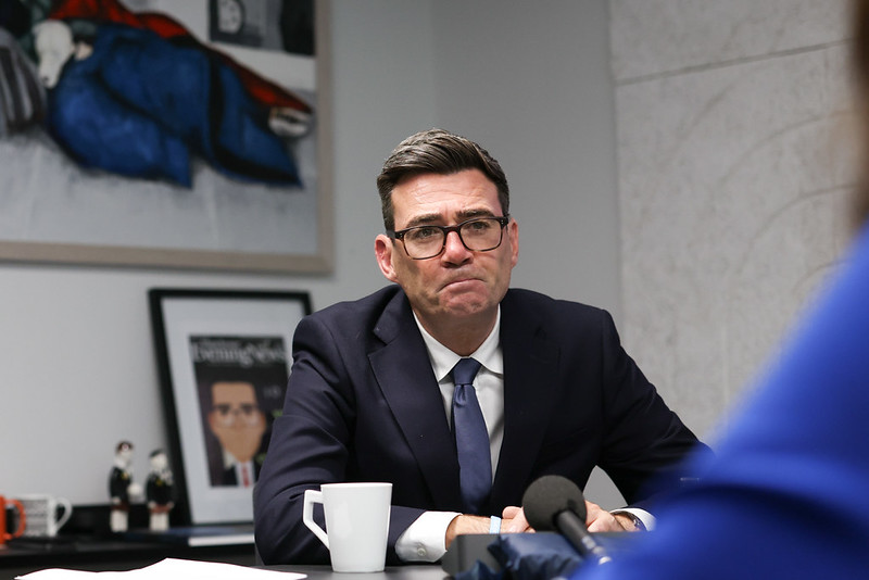 image shows the Prime Minister Andy Burnham sitting behind a desk at No 10 North. He is giving media interviews and there is a microphone pointed towards him