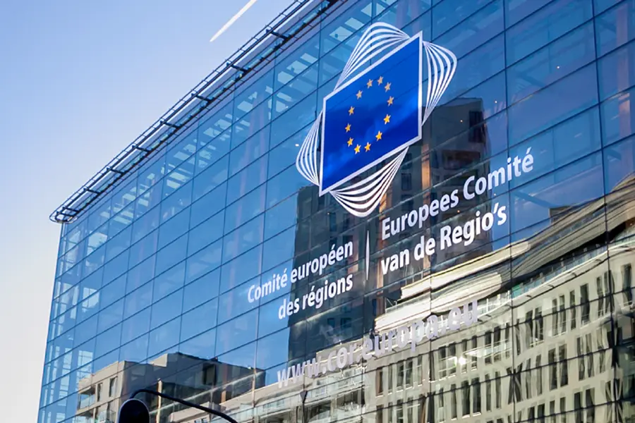 background l-r are the German, Union Jack and EU flags. image shows the exterior of a glass building with the European flag on the front and the words European Committee of the regions in French and Dutch.