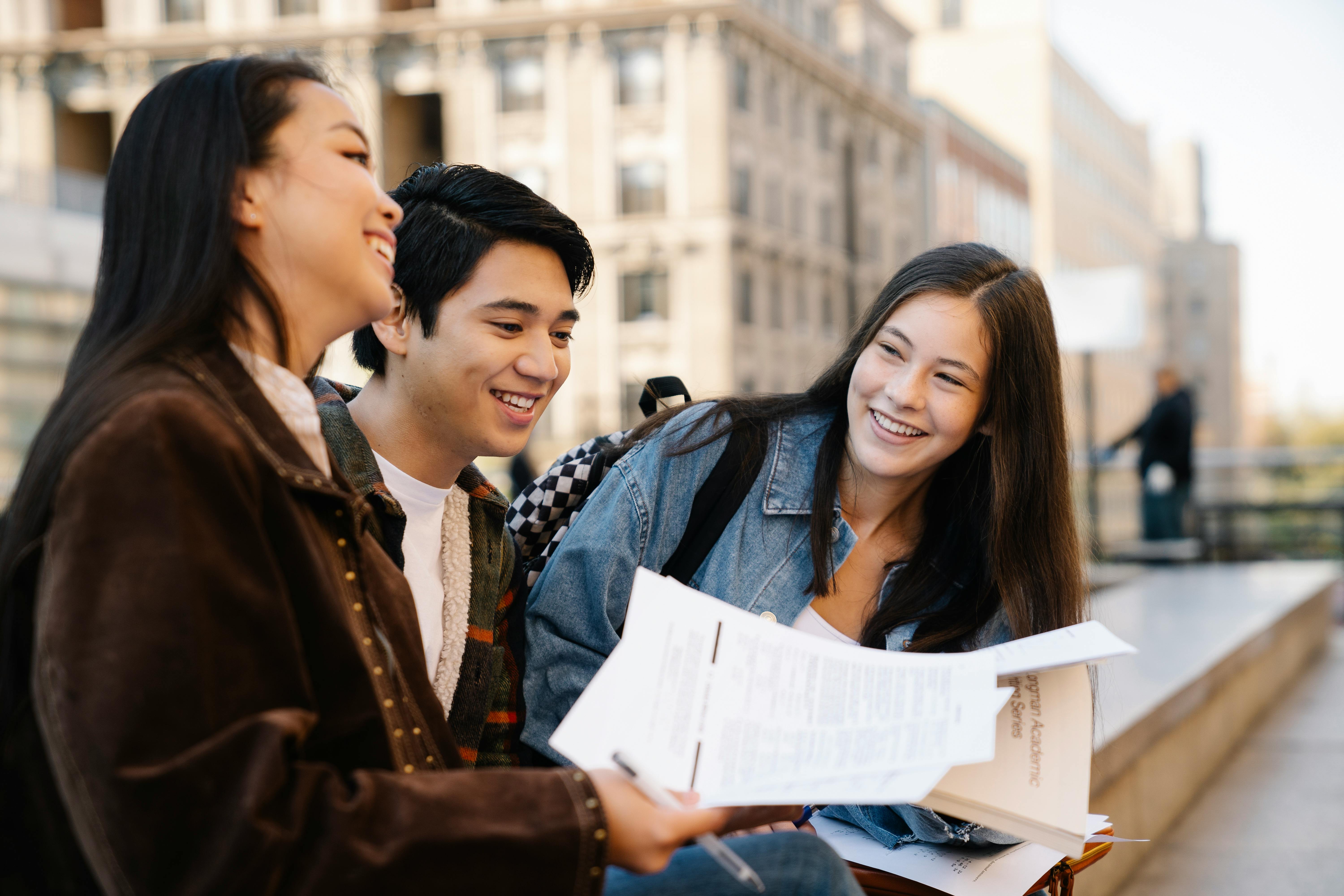 image shows yoing people holdinng papers and chatting while sitting on a concrete bench. 
