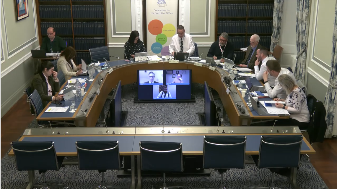 image shows Members of the Executive Office Committee sitting around the table in a Committee meeting room in Parliament Buildings. Slightly raised from the floor in front of the Chairperson are three screens which show the Directors of the Executive’s three overseas bureaux in Brussels, Beijing and Washington DC giving evidence to the Committee.   