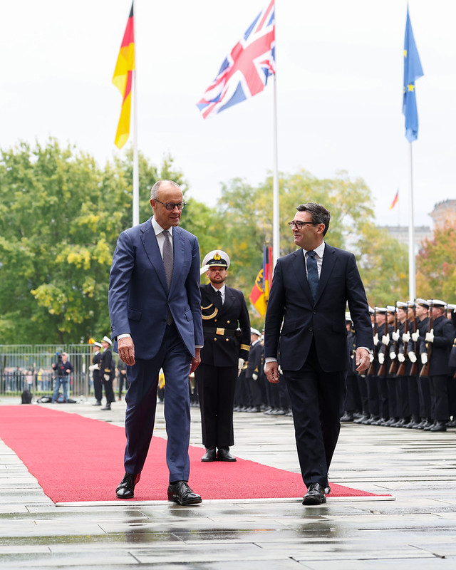 The image shows the UK Prime Minister on the right walking down a red carpet with Friedrich Merz, Chancellor of Germany at the Federal Chancellery in Berlin. In the background l-r are the German, Union Jack and EU flags.