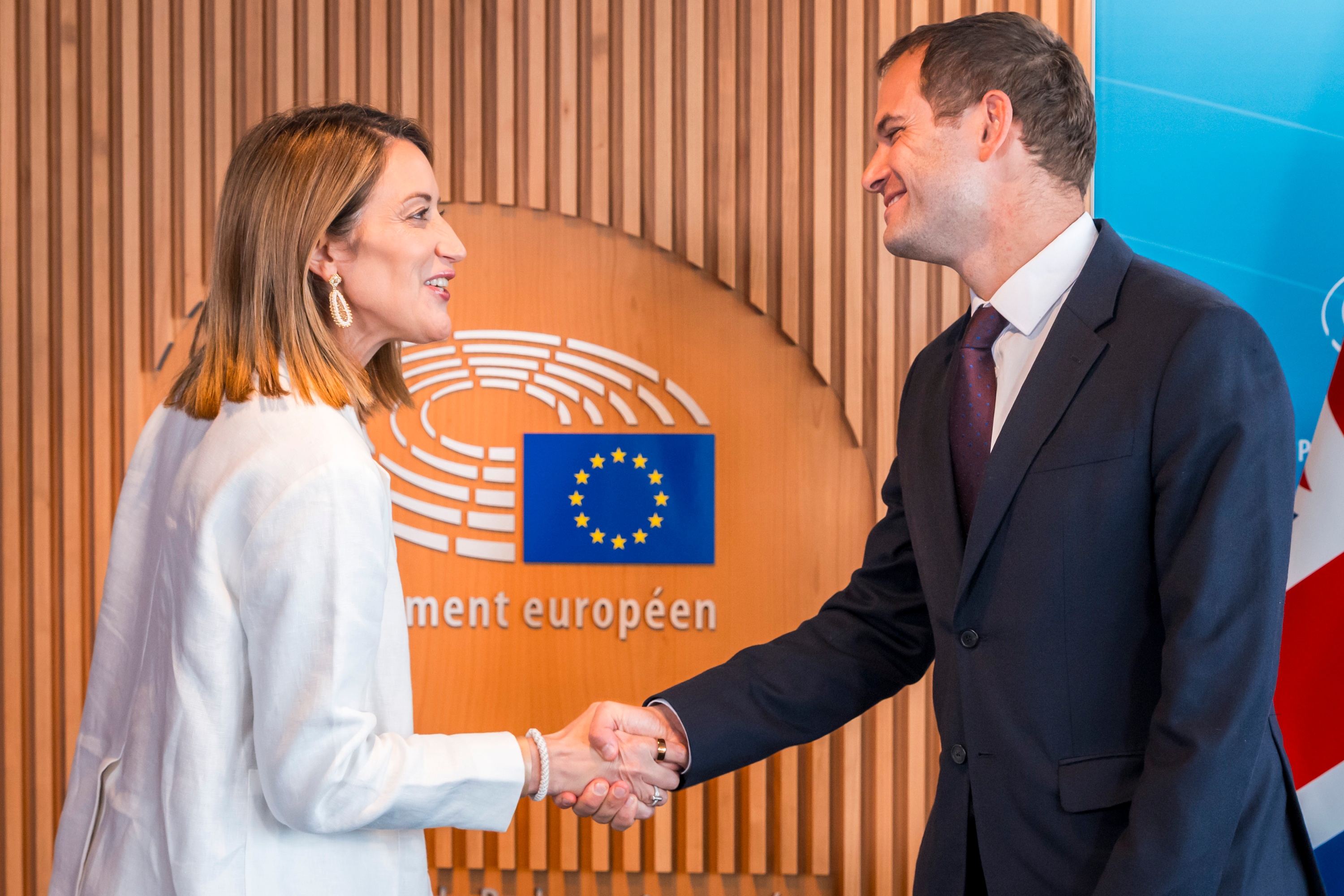 image shows President of the European Parliament, Roberta Metsola meets with Hamish Falconer, UK Minister for Intergovernmental Relations and European Relations. They are pictured shaking hands in front of an image of the seating in the EU Parliament and an EU flag.