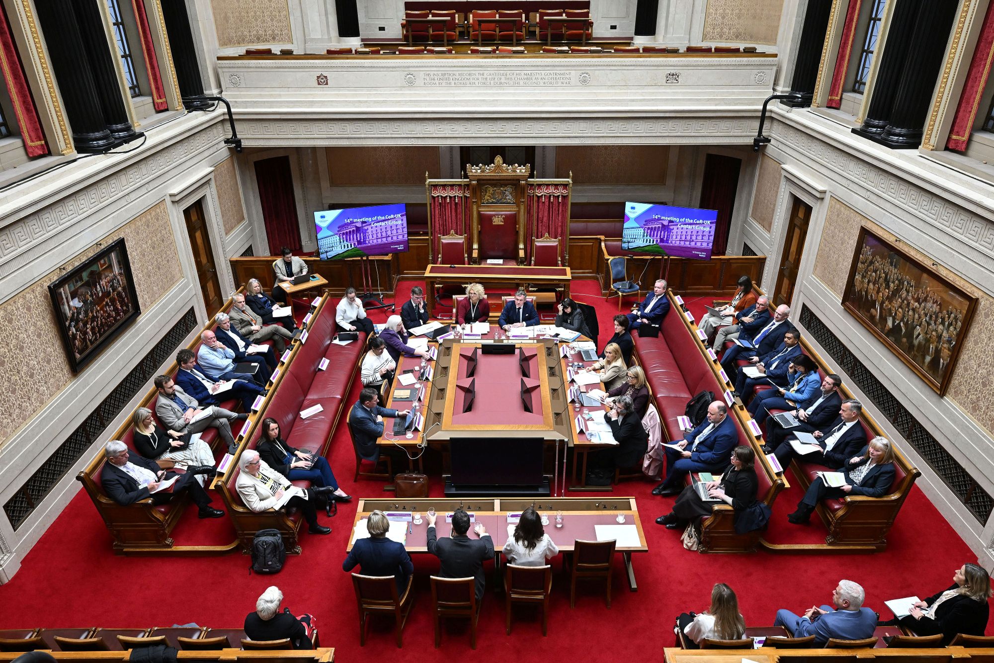 Image shows members of the Committee of the Regions (CoR) – UK Contact Group pictured at a meeting in the Senate Chamber in Parliament Buildings. Members are seated at the top and on both sides of a long square table in the centre of the room, other Members are seated in red coloured benches on either side. The photograph is taken from the Senate Chamber gallery looking down into the room.