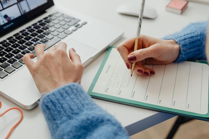 image shows arms resting on a desk. The left hand is typing on a laptop and the right hand is writing in a planner. 
