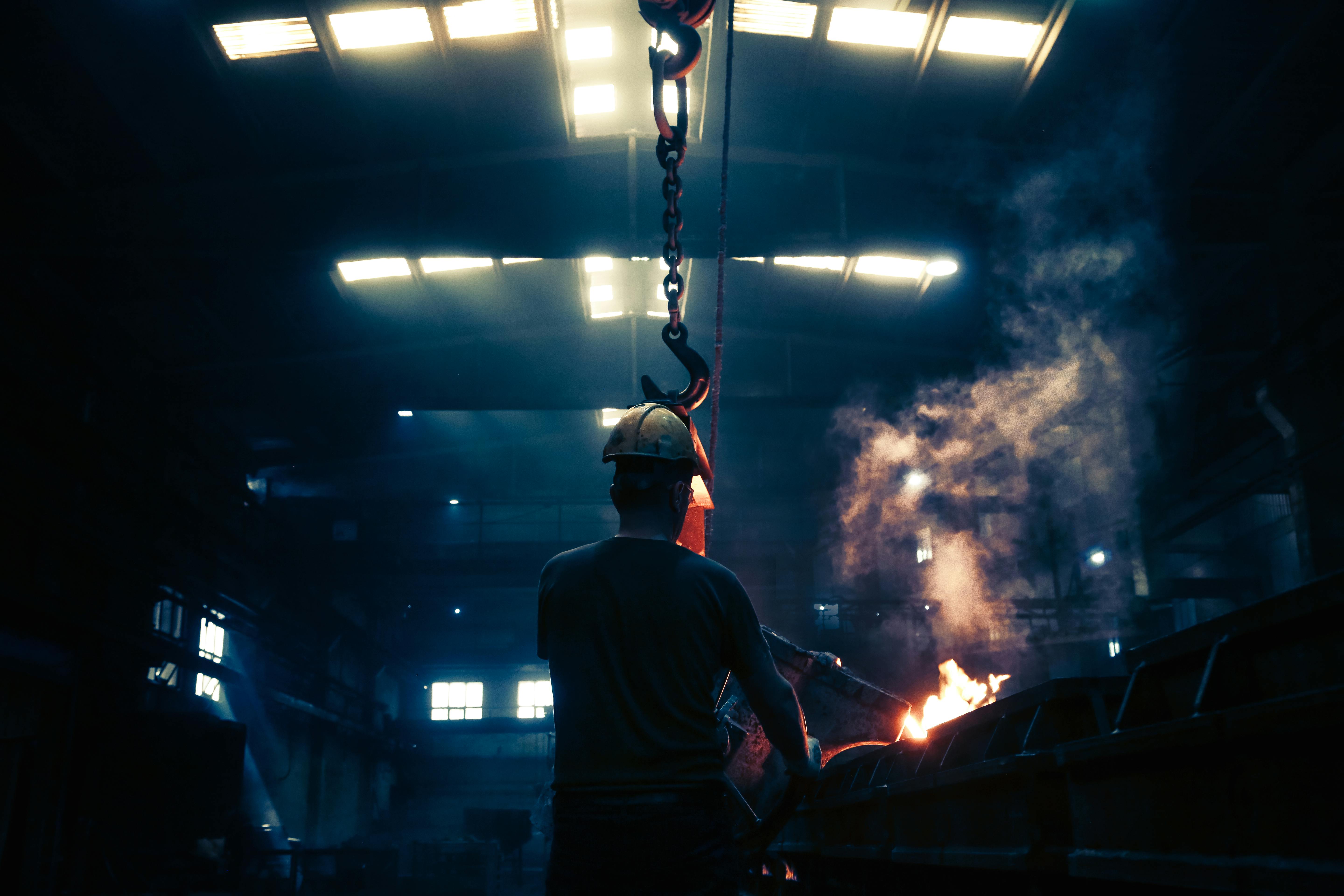 image shows a person in a hard hat working in heavy industry - his back is to the camera and the photograph is dark apart from sparks coming from the equipment to his right.  