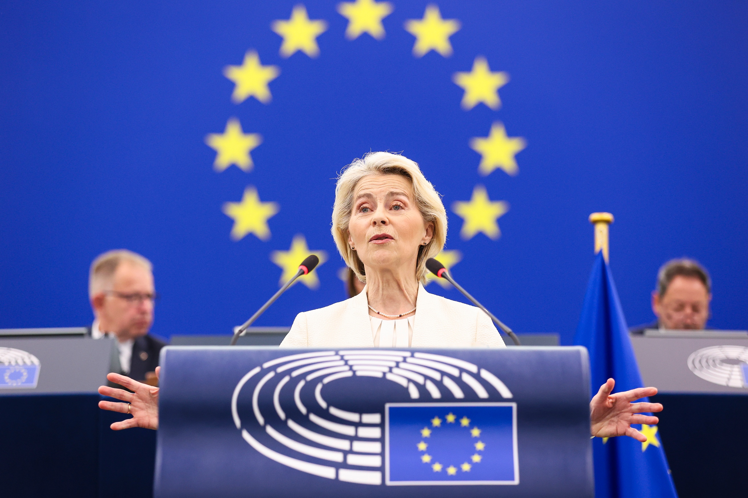 image shows the President of the European Commission, Ursula von der Leyen giving the State of the Union address to MEPs. She is standing against a blue background with a circle of gold stars just behind her head
