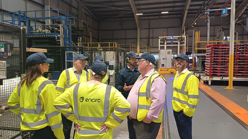 Committee members being shown around a glass factory by representatives of Encirc. Members are identified in the paragraph below. Each member is wearing hi-viz protective equipment, hard hats and goggles during the tour. We see a tour guide describing the factory to the members.