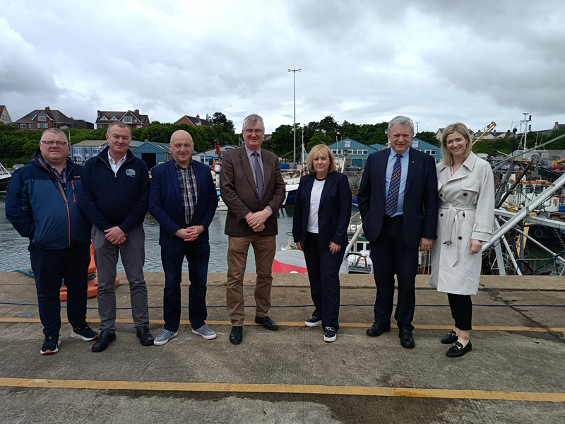 Members of the Agriculture Committee with a representative of ANIFPO on the water's edge of Kilkeel Harbour. Details of the individuals in the picture are available in the caption below. In the background we can see boats and boathouses. It appears to be a busy harbour.