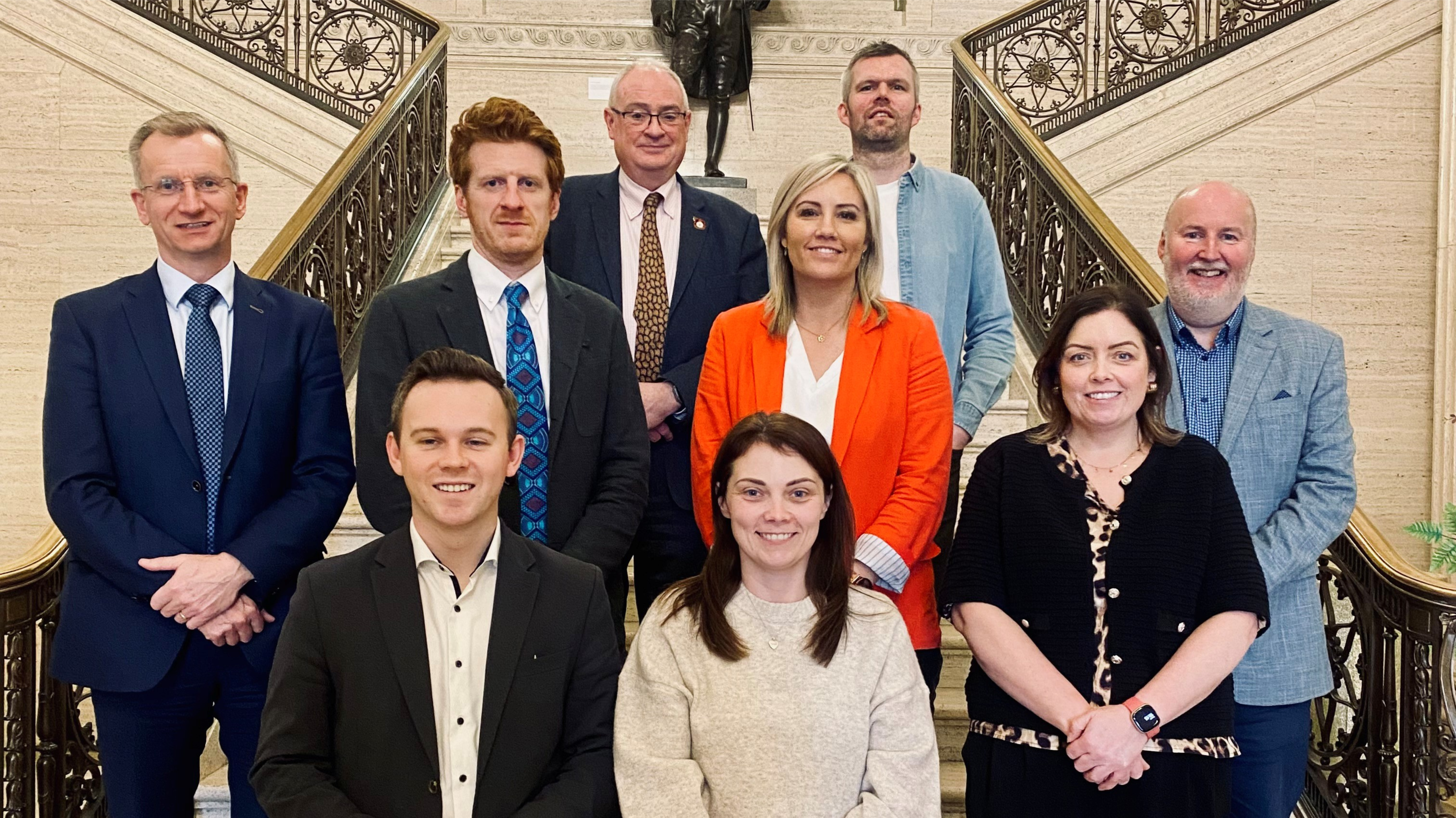 Image shows members of the Finance committee on the steps inside the Great Hall at Parliament Buildings. From right to left from the back row: Steve Aiken, Gerry Carroll, Brian Kingston, Matthew O'Toole, Diane Forsythe, Harry Harvey, Eoin Tennyson, Jemma Dolan and Deirdre Hargey.