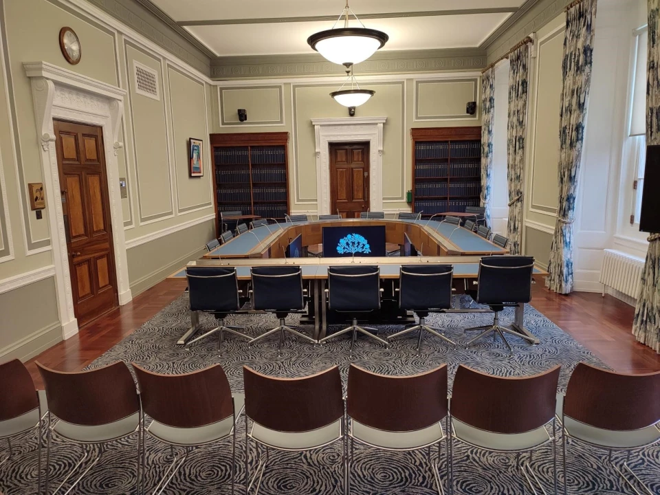 One of the Assembly's Committee rooms. The meeting table is horse-shoe shaped. The 'arc' of the horseshoe is where the Committee chairperson sits with the other members of the Committee sitting around the sides. Witnesses sit at the 'base' of the horseshoe, facing the Committee. There is a viewing gallery behind the table which is made up of empty seats which members of the public are welcome to sit in when the Committee is in session.