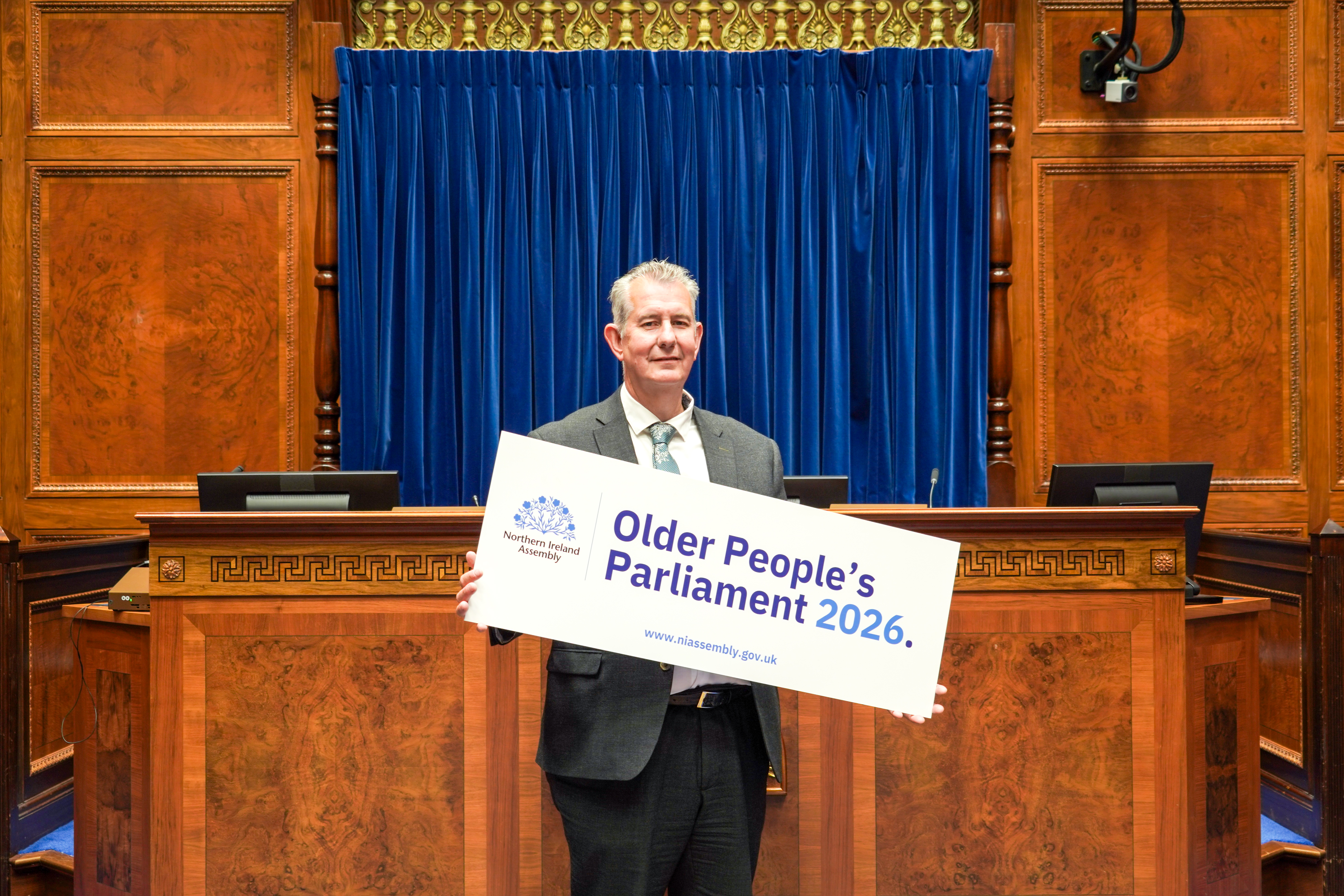 Assembly Speaker, Edwin Poots MLA standing on the Chamber floor holding a sign informing people of the upcoming Older People's Parliament happening in October 2026 Assembly Speaker, Edwin Poots MLA standing on the Chamber floor holding a sign informing people of the upcoming Older People's Parliament happening in October 2026